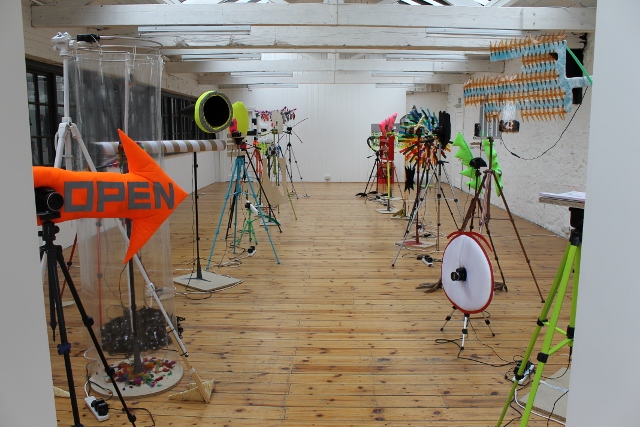 A collection of sculptures on a wooden floored gallery space. In the foreground one of them has a n orange sign that is arrow shaped it says 'OPEN'.