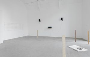 A wide white cube gallery space. There are three small black objects on the far wall. A light timber pole in the foreground.