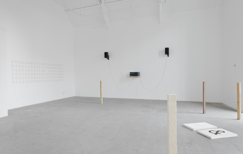A wide white cube gallery space. There are three small black objects on the far wall. A light timber pole in the foreground.