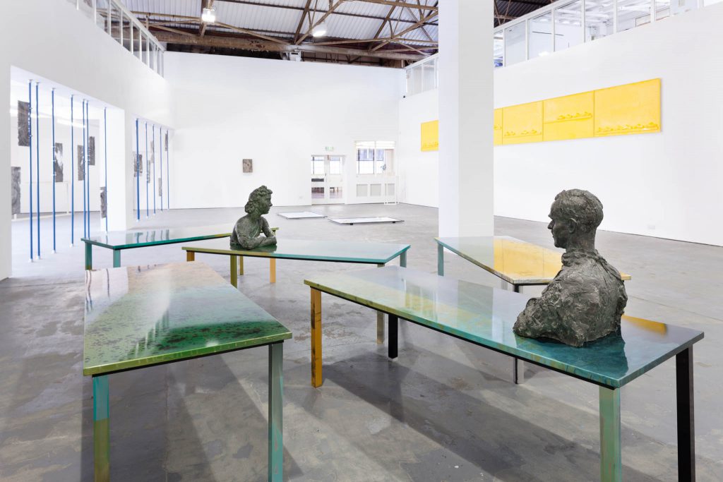 Two green topped tables in a gallery space. Each has a bronze bust on. There are a number of pieces on the large white walls behind.