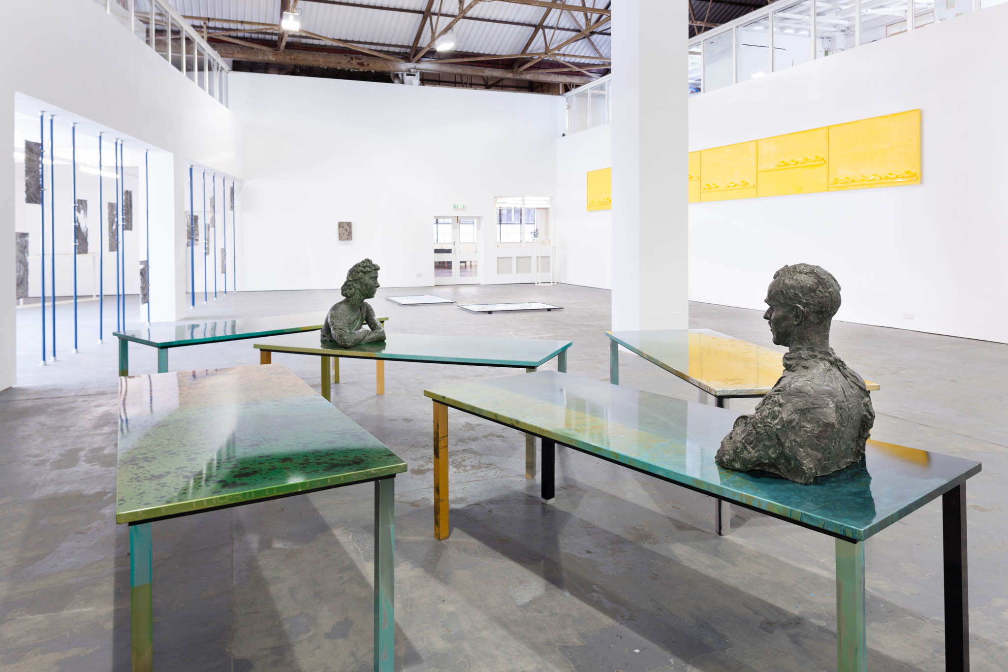 Two green topped tables in a gallery space. Each has a bronze bust on. There are a number of pieces on the large white walls behind.