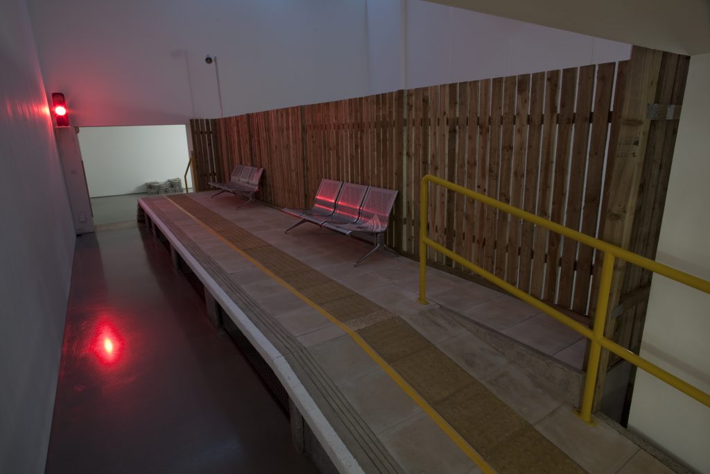 A train station platform brought into a gallery. It features the concrete and tarmac platform itself, red lights to warn about approaching trains, grey benches, yellow handrails and a tall wooden fence.