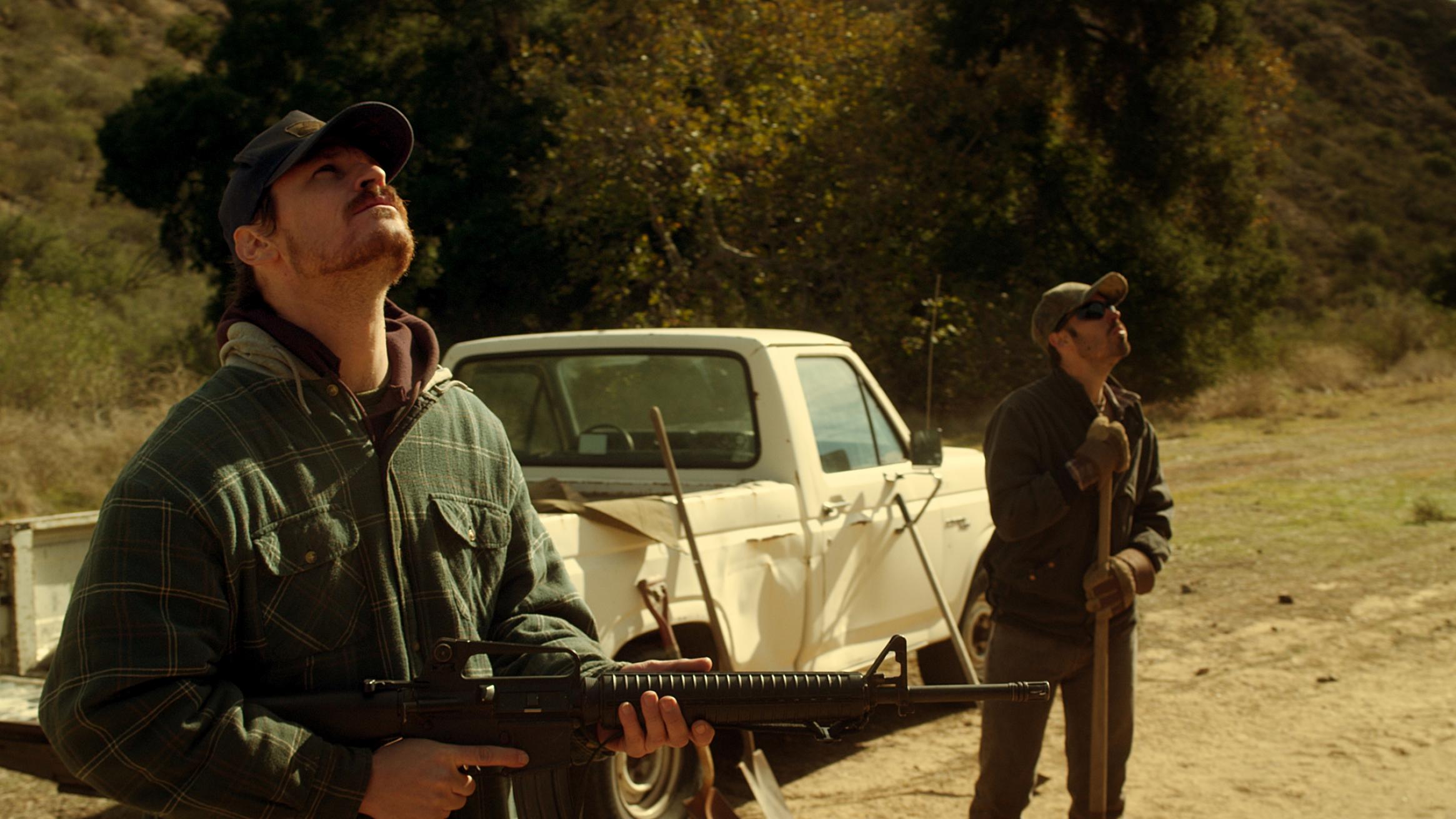 A photograph of two men in military uniform stood by a white car on a dusty road. The man in the foreground is looking up.