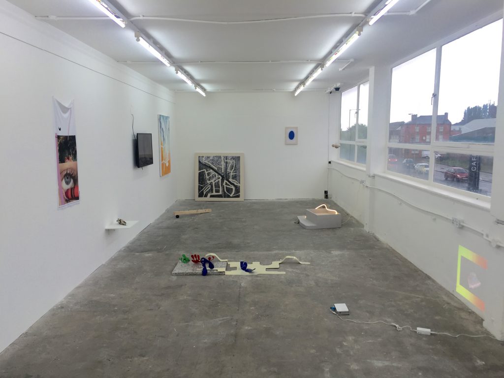 Long white gallery space flanked on the right with a long window. Art objects line the left-hand and back wall, and lie dotted around on the floor