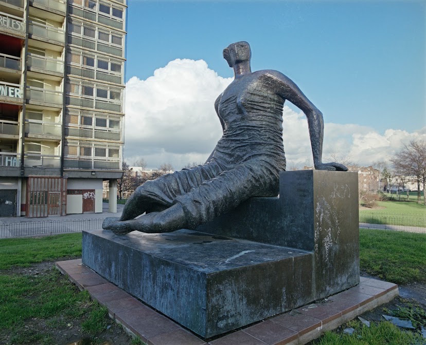 A bronze sculpture of a seated woman. Her figure is abstract and angular, and she sits upon a set of bronze steps.