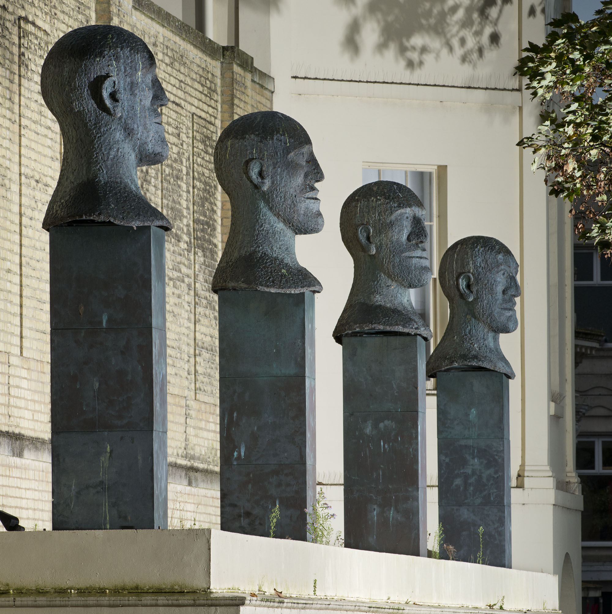A series of four sculptural heads on plinths. The heads are shown in profile and all look in the same direction. They are somewhat masculine but have few other defining features.