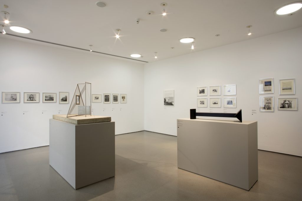 A view of a white wall gallery. Framed works are hung on the walls and two plinths hold sculpture in the centre of the room.