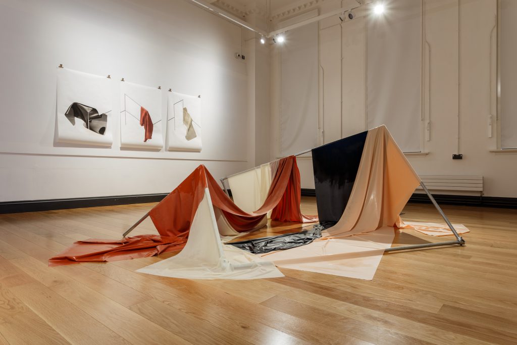 View of a gallery with a large fabric installation in the centre. It appears to be made up of different sheets arranged into geometric shapes. The colours are peach, orange, beige and black.