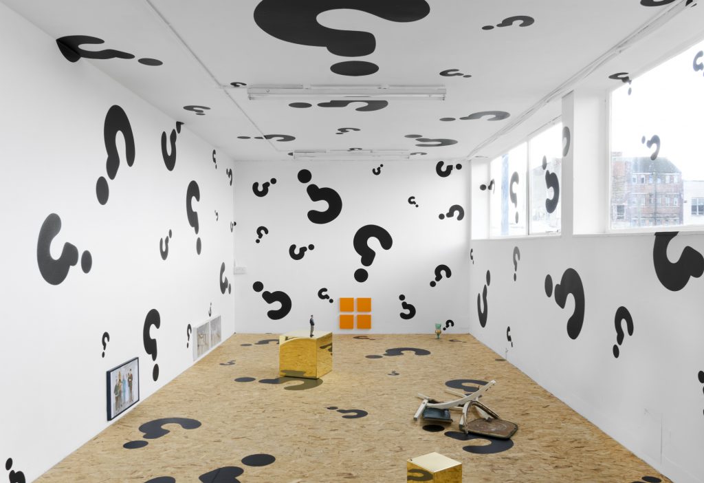 A gallery space with white walls and wooden floor. All surfaces of the room, including the windows, are covered in black question marks.