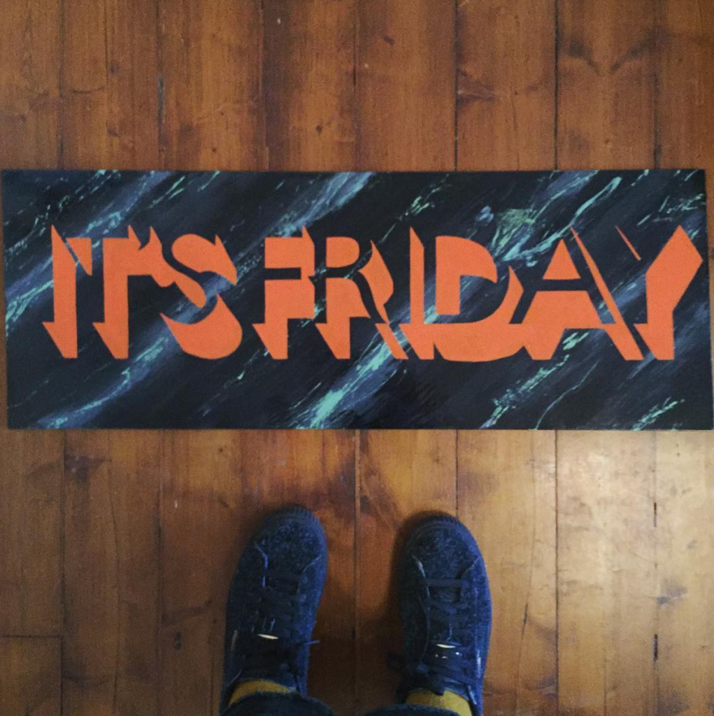A sign reading 'It's Friday' photographed on a wooden floor, the photographer's feet are visible next to the sign.