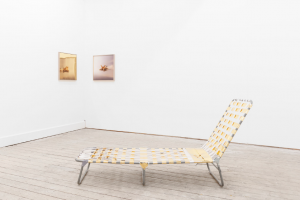 A sun lounger chair in a gallery, with two works behind it on the wall.