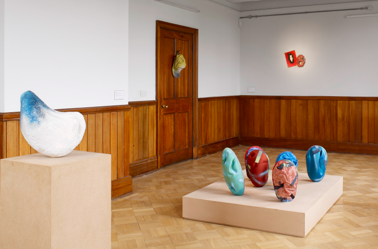 A gallery with wood panelling and white walls. Several colourful sculptures rest on a plinth close to the floor.