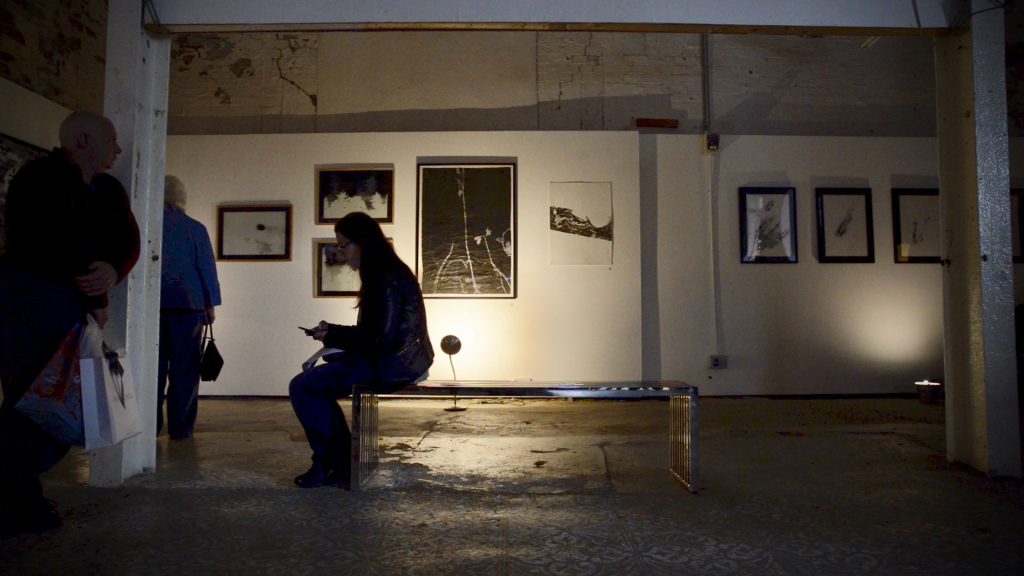 A person sits on a bench in a darkened gallery space. Works on the wall are just visible behind them.