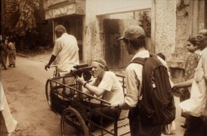 A sepia photograph of three people. Two are walking, one is in the trailer of a bike holding a camera to film.