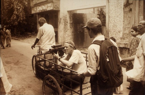 A sepia photograph of three people. Two are walking, one is in the trailer of a bike holding a camera to film.