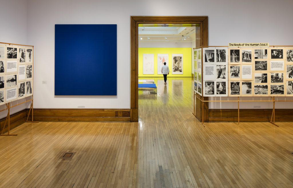 A gallery with white walls and wooden floor. In the first room boards are arranged with black and white images and text. The title reads 'The needs of the human soul'. A large blue painting hangs next to them. Visible in the next room are yellow walls and more framed works.