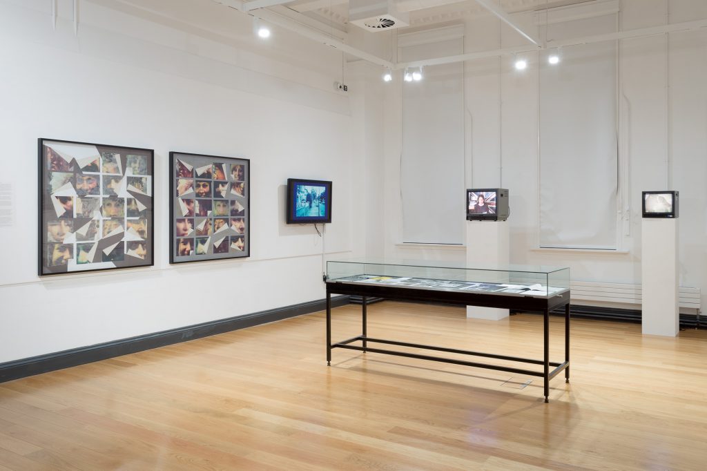 View of a gallery space with white walls and pale wooden floor. There are several works visible: a vitrine with black legs and several small TVs screening artists' films.