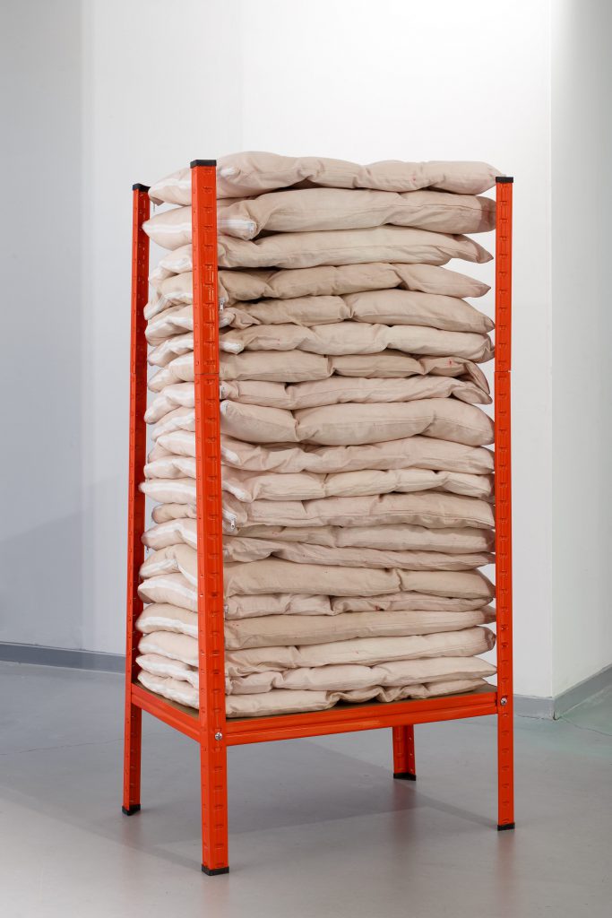 A red metal shelving unit containing a stack of beige and peach coloured soft sculptures. They appear like pillows or folded quilts.