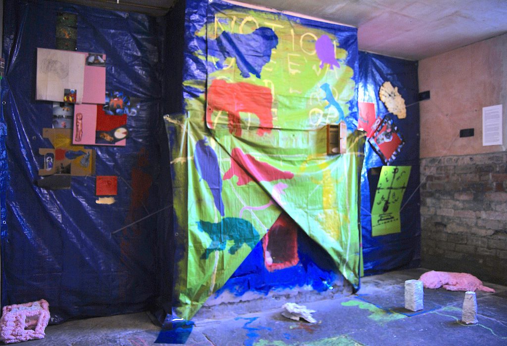 A large fabric wall covering in shades of green, blue and red. The shapes of animals can be made out. On the floor in front are small sculptures, and other works can be seen hung on the walls of the room.