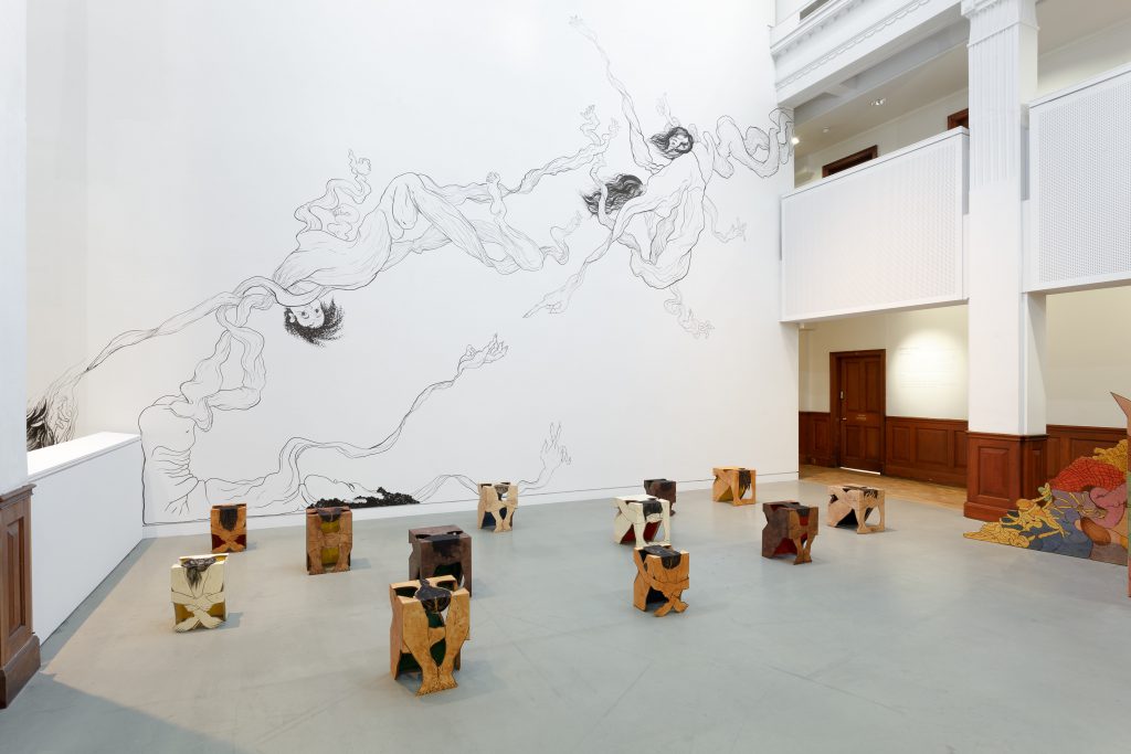 The atrium of a gallery. Arranged on the floor are a series of wooden sculptures depicting women seated with their legs drawn up to their chests and heads down. They look like a series of wooden stools or cubes. On the wall behind is a large drawing showing bodies stretched out to become tentacles, clouds and drifts of smoke.