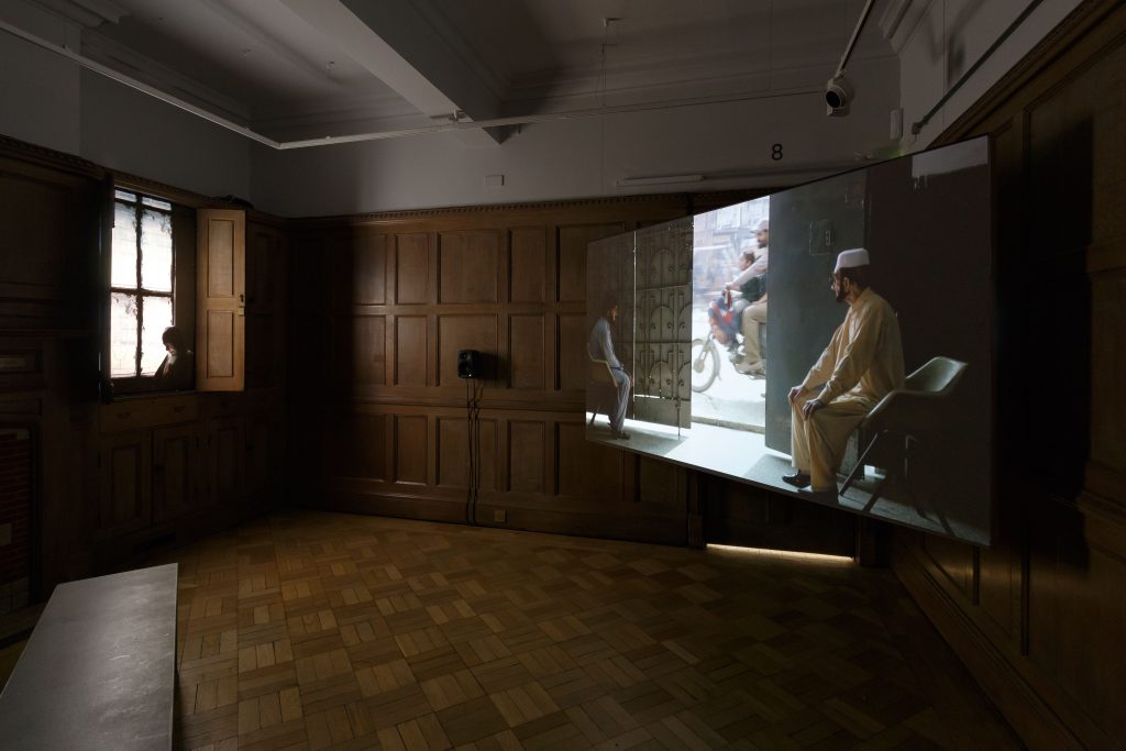 A darkened gallery with an artist's film being screened. The image still shows a seated man and an open doorway to a busy street.