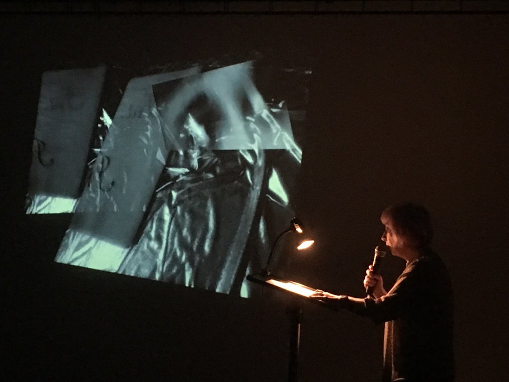 A person stands at a lectern, with a small light shining on their pieces of paper. They hold the microphone up to read. In the background there is an abstract projected image in shades of grey, green and silver.