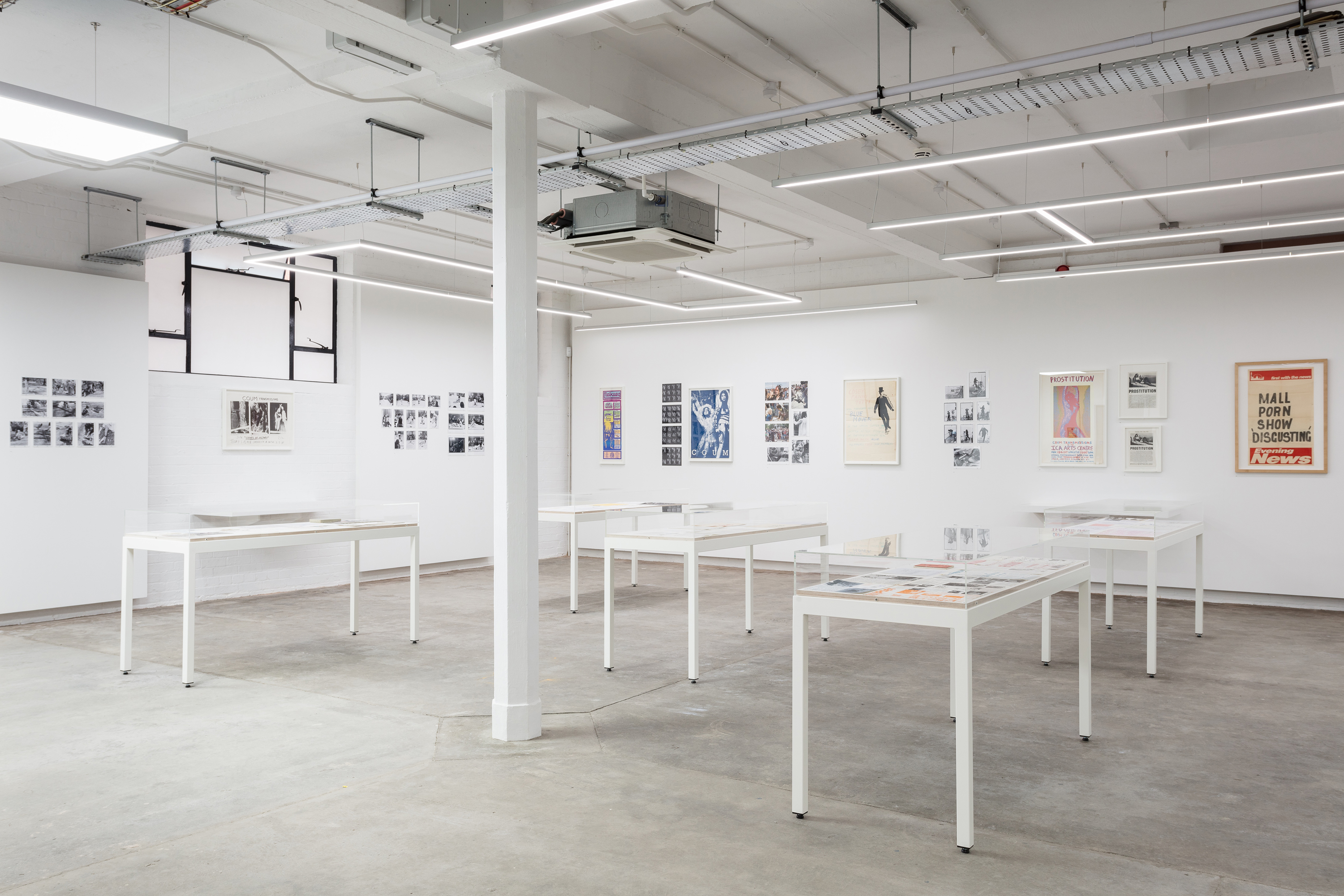 View of a white wall gallery with grey floor and white pillars. In the space there is lots of archival material presented in small groups on the wall, and in white plinths.