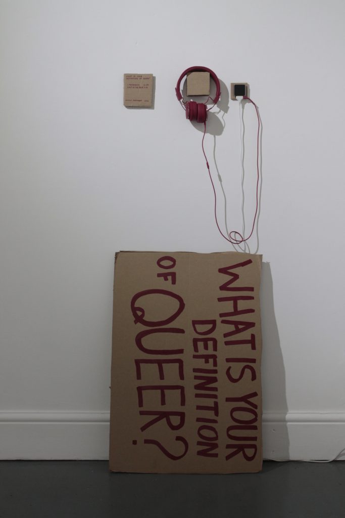 A cardboard sign on its side that reads: 'what is your definition of queer'. Above it are some red headphones and a small cardboard label.