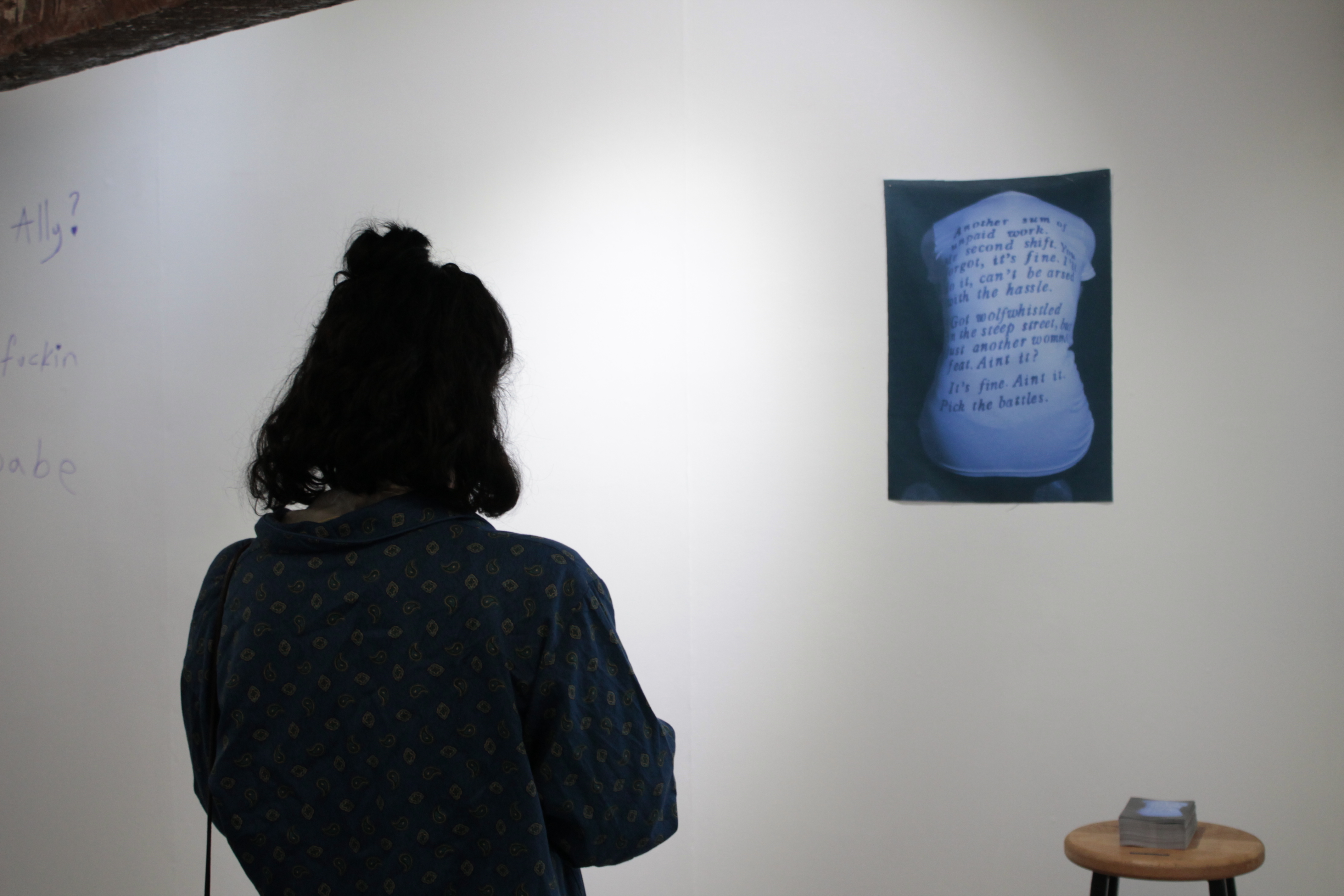 A person looks at an artwork mounted on the gallery wall. The work shows a person's back, overwritten with text.