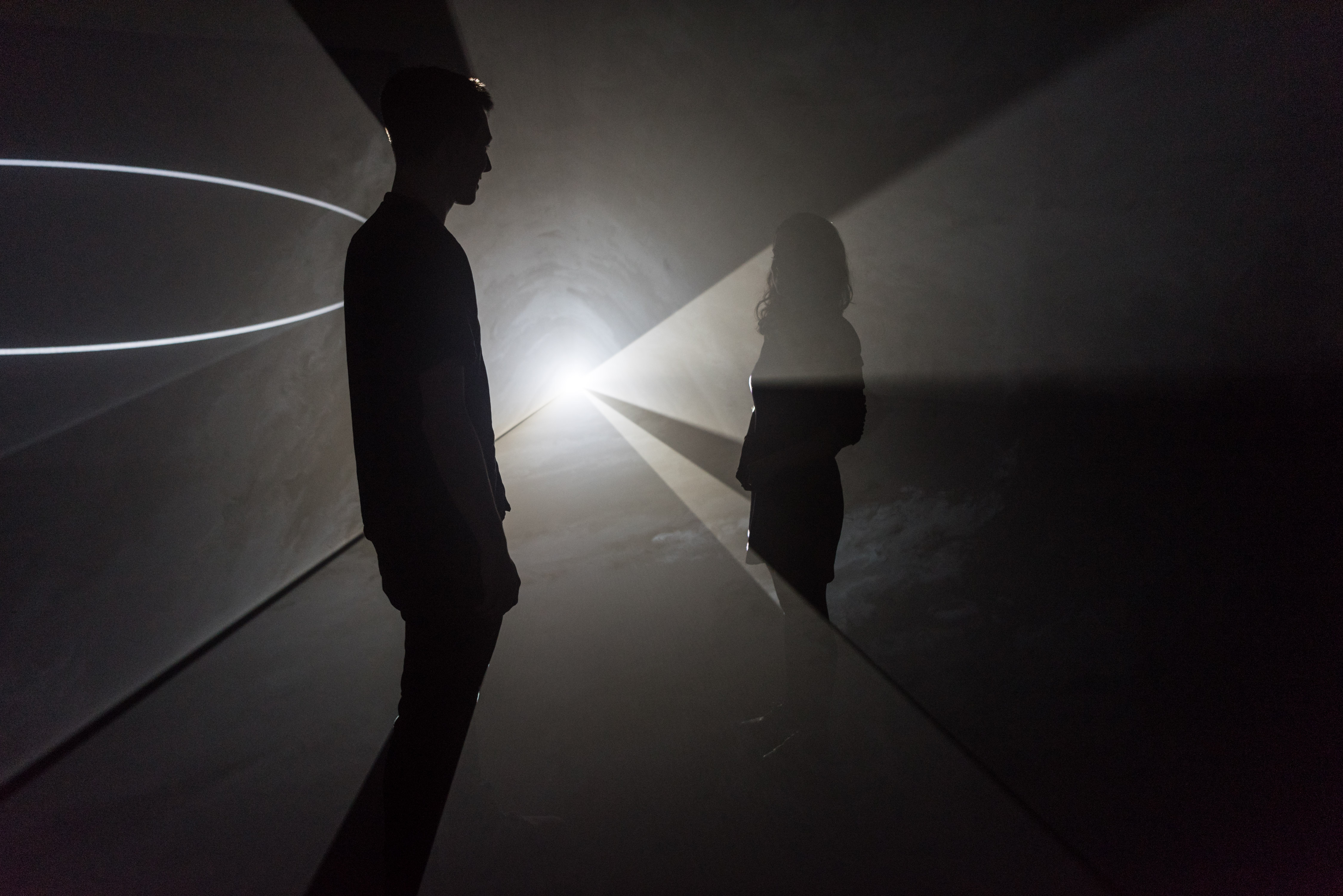 Two people stand in a darkened room, with a white light shining through them.