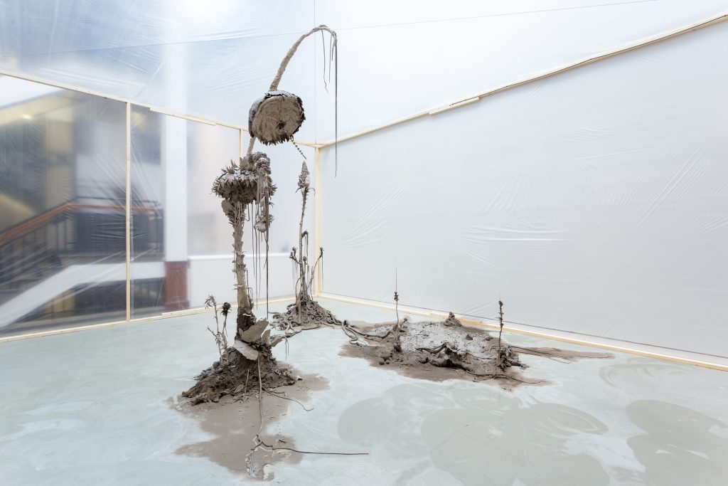 A sculpture made of clay and armatures. It gives a sense of growth and decay, recalling soil and plants as well as post-industrial landscapes. Around it are simple stud walls covered in clear plastic.