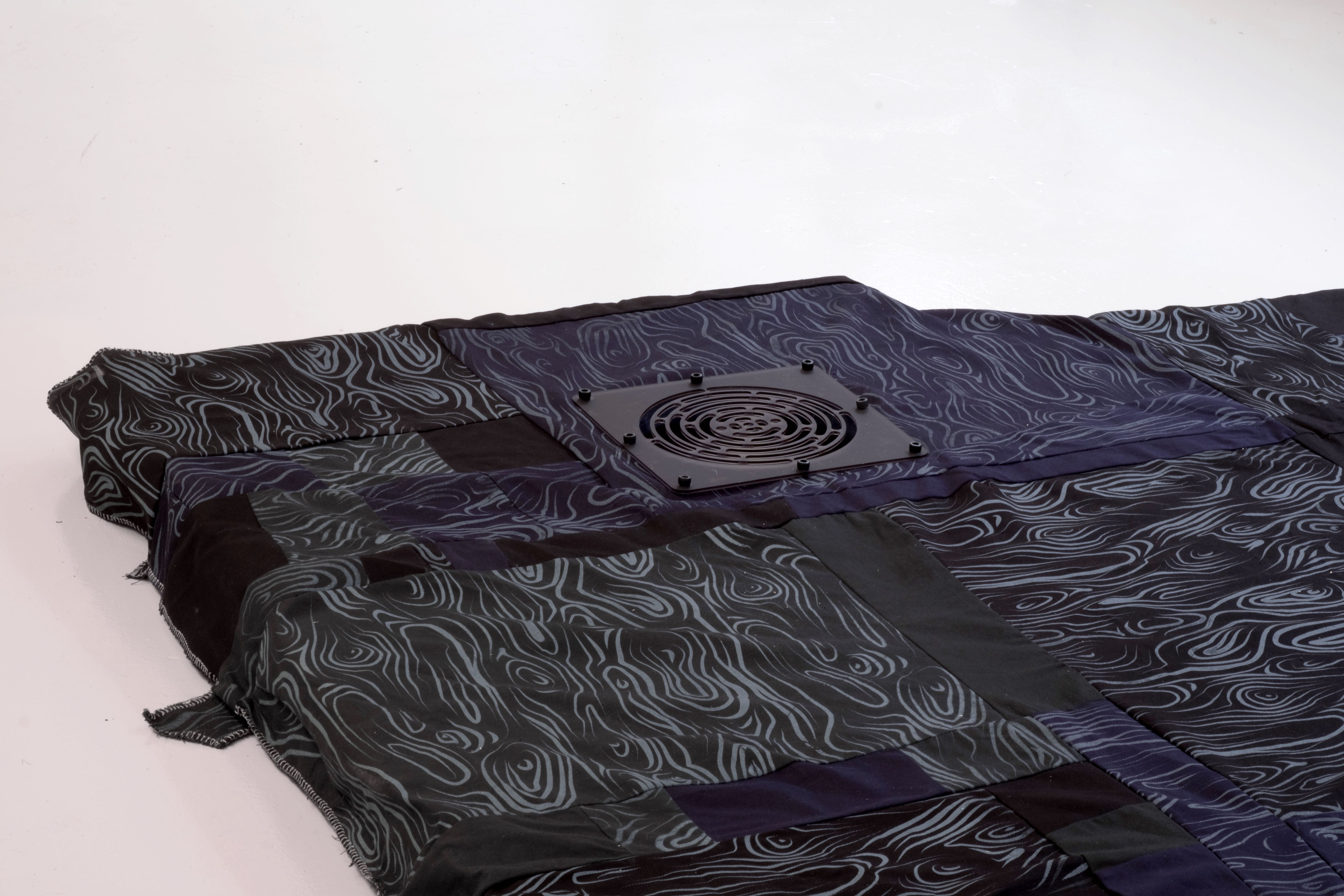 A black fabric object, with a black grate inlaid. There are many patterns across the surface of the work.