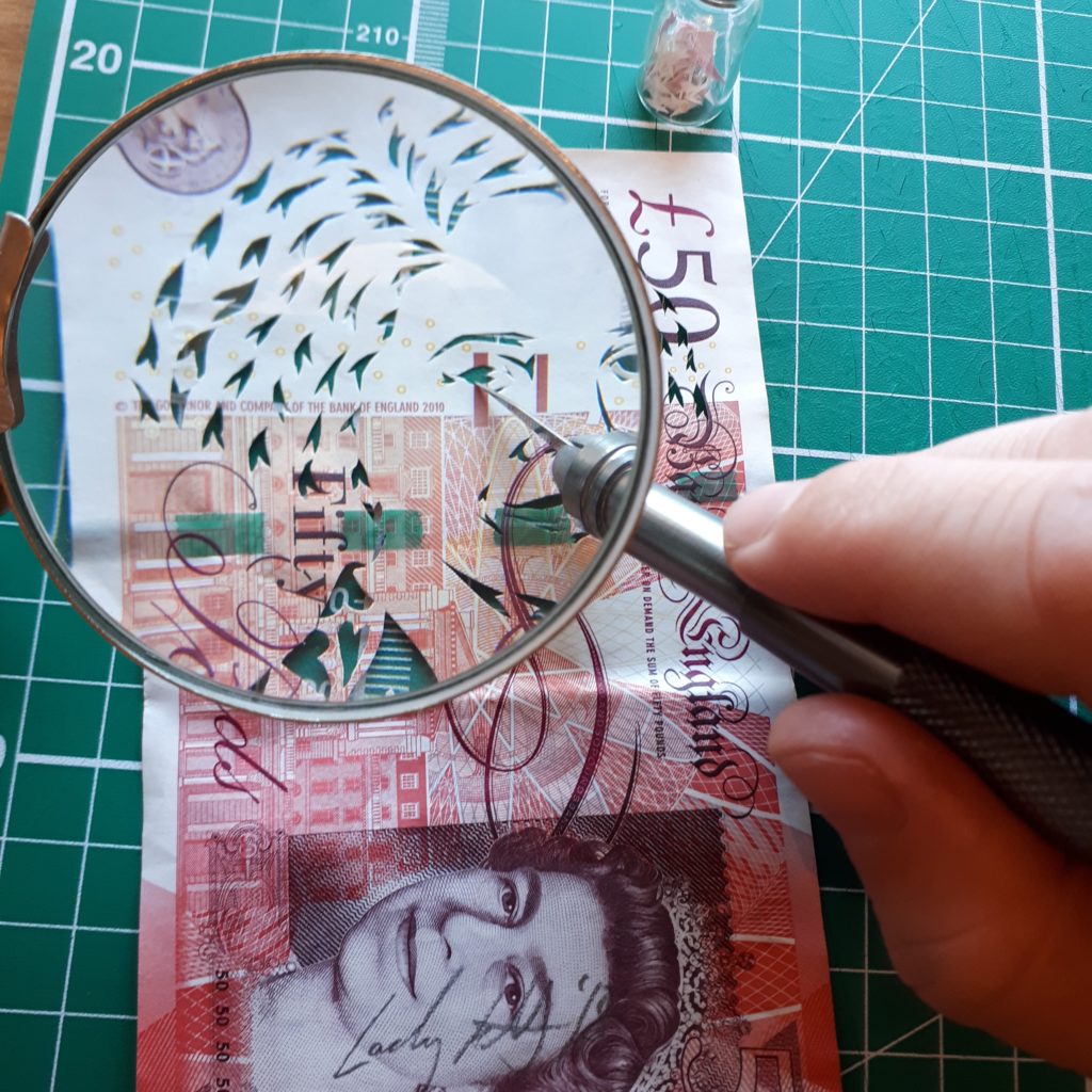A close up of a scalpel cutting a drawing of a face into a 50 pound note.