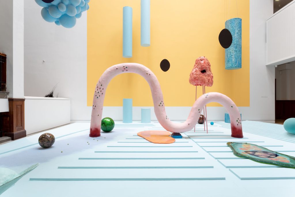 A view of a colourful and complex art installation. There are lots of sculptural objects and shapes in shades of pale pink, blue, green and yellow.