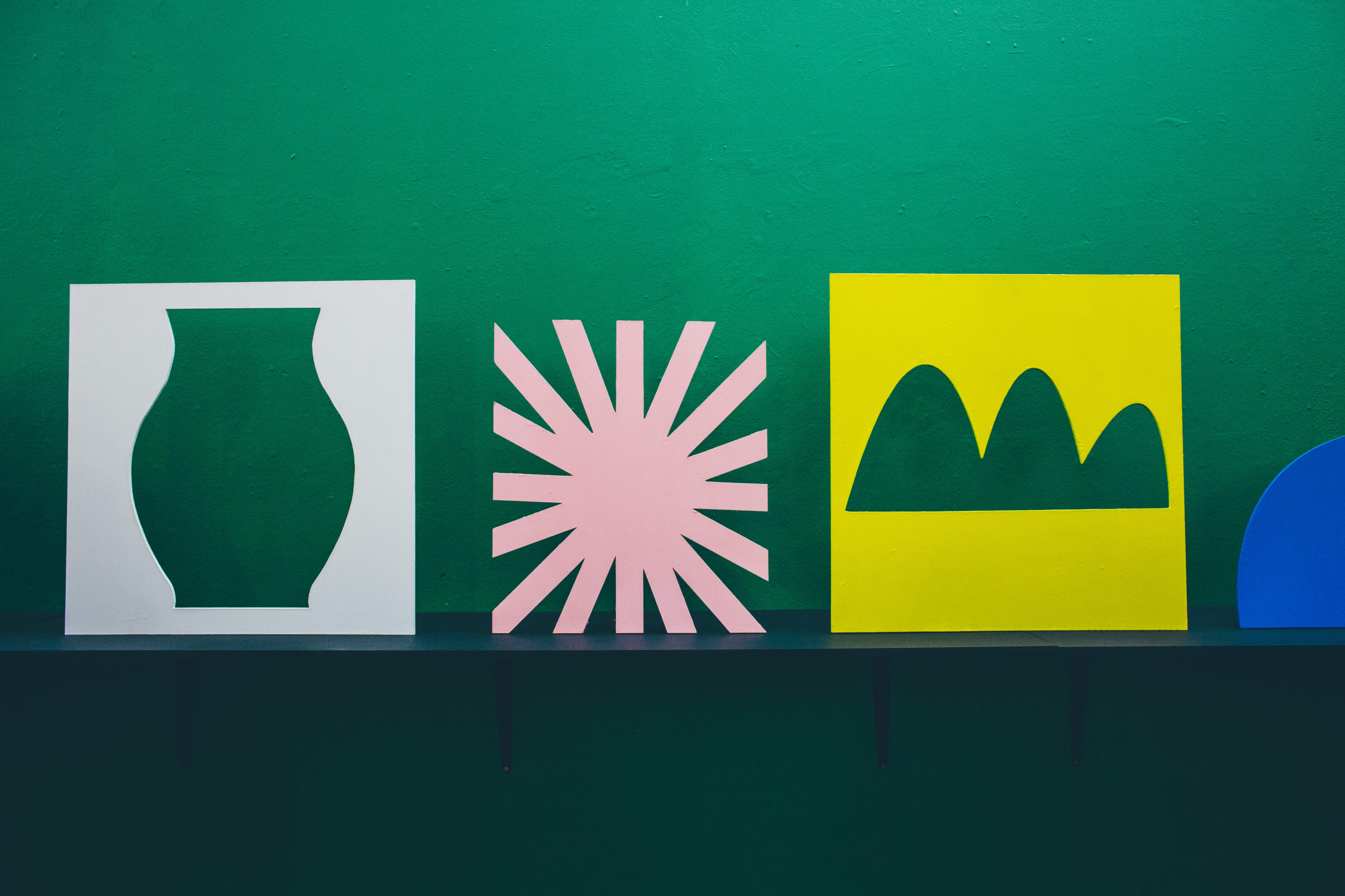 An image of three cut-out artworks against a dark green wall. They depict a vase, a star or sun, and a trio of hills.