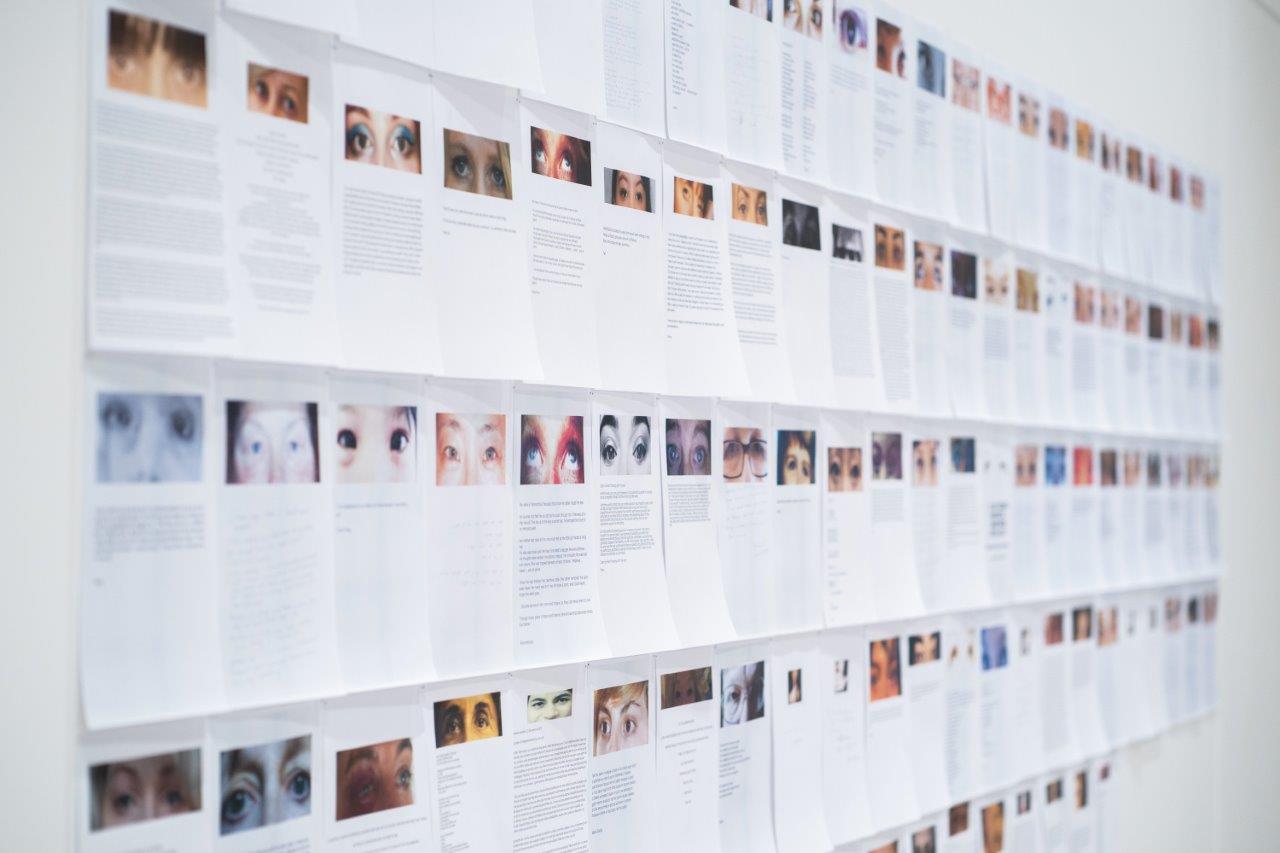 A series of printed paged pinned to a gallery wall. Each page has an image, mostly of women's eyes, and blocks of text.
