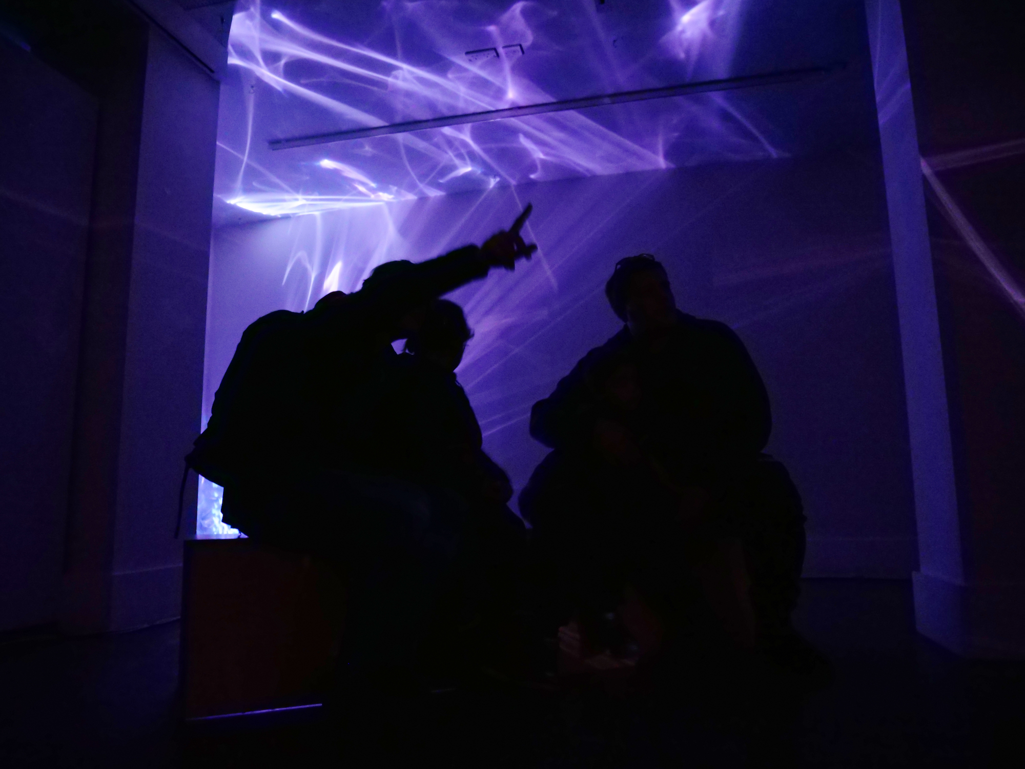 Three silhouetted bodies in a room lit by purple and white shapes and patterns.