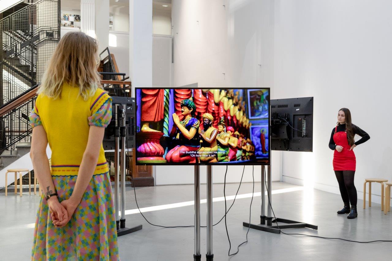 Two people stand in a white wall gallery, they are watching artists' films on TV screens throughout the space.