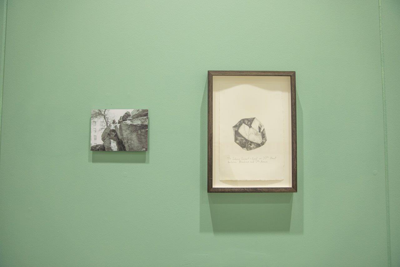 A green gallery wall featuring two artworks. On the left, a black and white image of rockface with a building in the background. On the right a drawing of a rock or mineral.