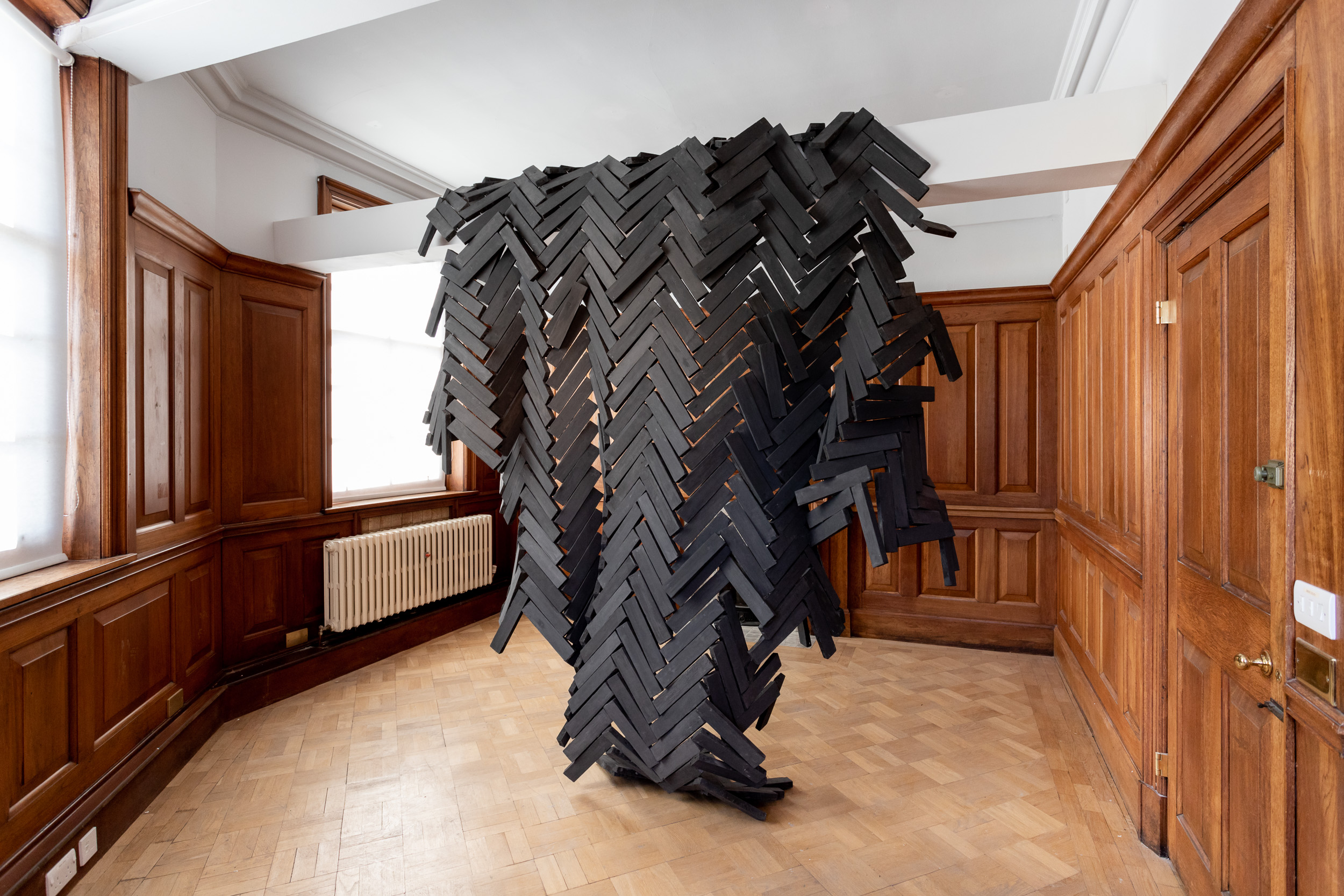 A sculpture in a gallery room with wooden floor and wood panelled walls. The work is made up of small pieces of black wood, arranged in a geometric and tessellating pattern in three dimensions.
