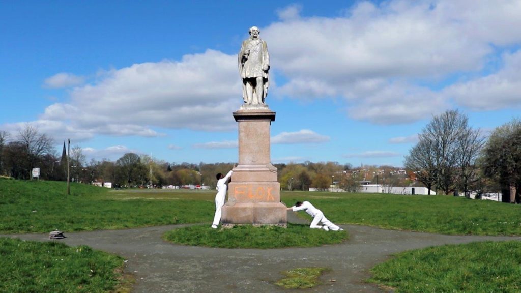 A park with a large monument. Two people dressed in all white push and pull at the sides of the statue and plinth.