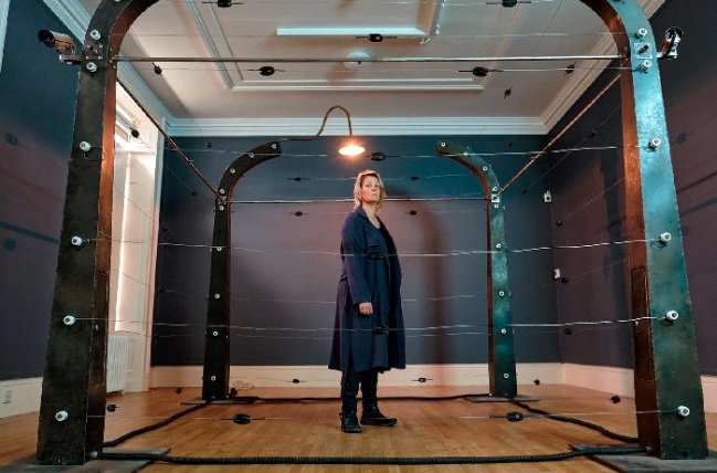 A white woman with blonde hair stands in the middle of an artwork made up of a square of electric fencing.