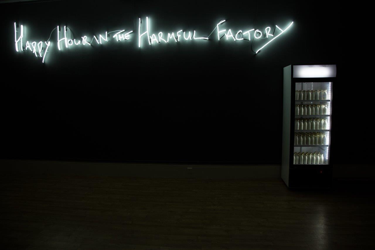 A neon white sign that reads: 'Happy Hour in the Harmful Factory'. Next to it is a commercial fridge containing bottles of milk, lit up from within.