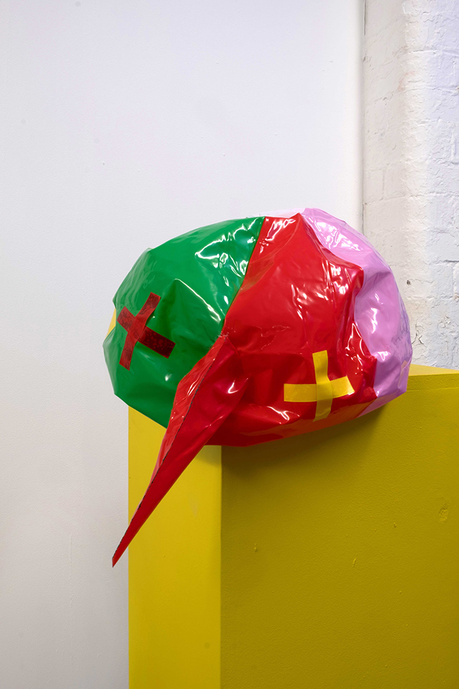 A sculpture made of different coloured plastics in the shape of a ball. It looks like a face, with crosses for eyes and a long pointed nose. It rests on a yellow plinth.