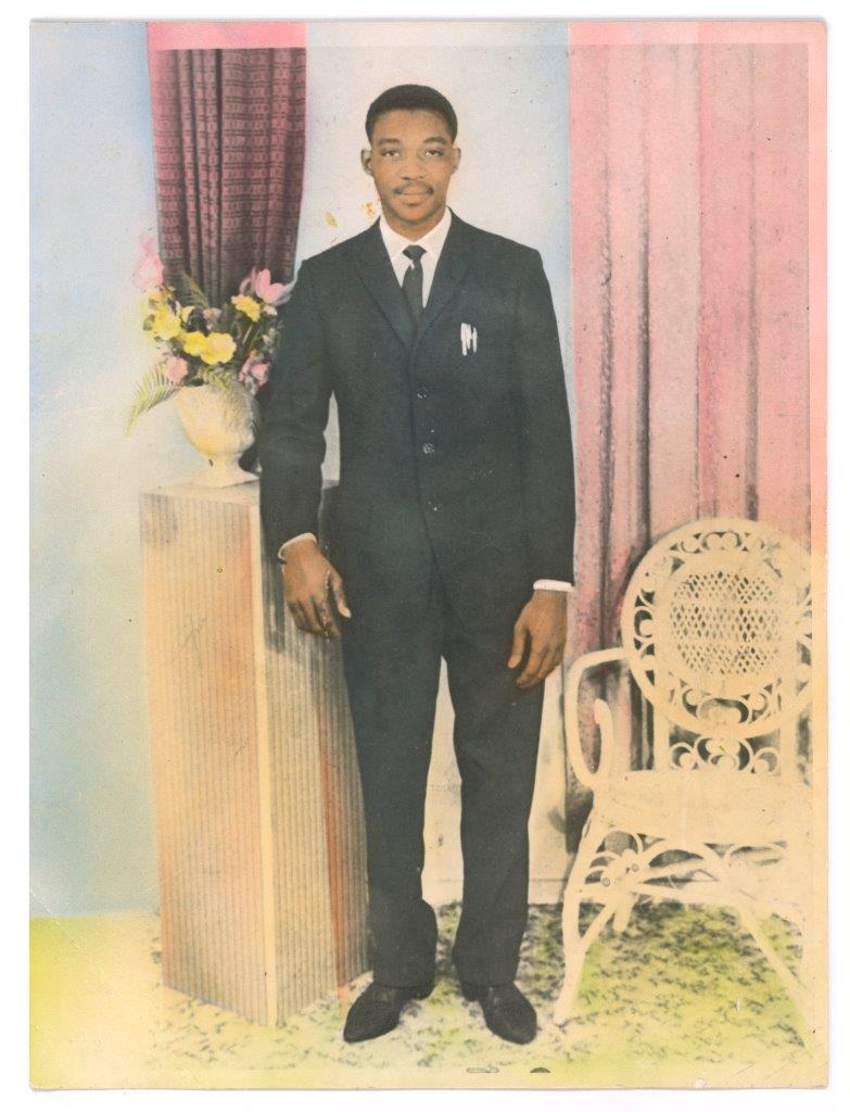 A studio portrait of a black man in a suit, he stands smiling at the camera.