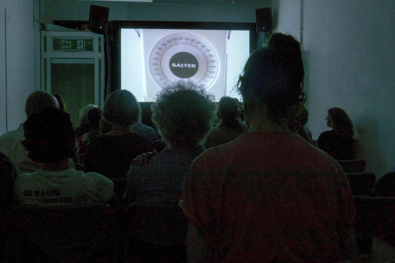 A photograph of a film screening. The audience silhouette's fill the room, on screen is a still showing a Salter weighing scale.
