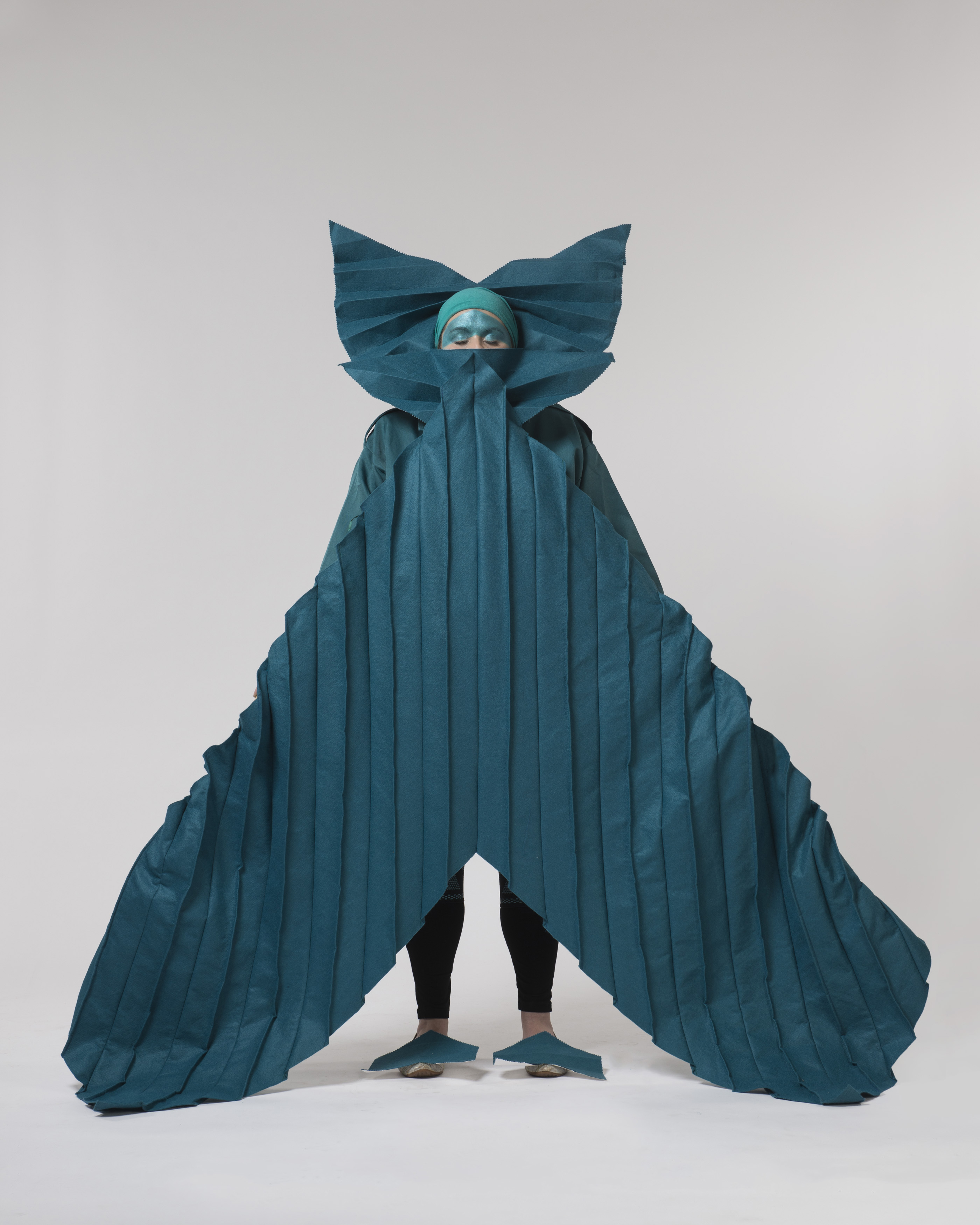 A person standing against a plain background. They are wearing a huge turquoise costume and mask, which extends their shape to appear like an arrowhead.