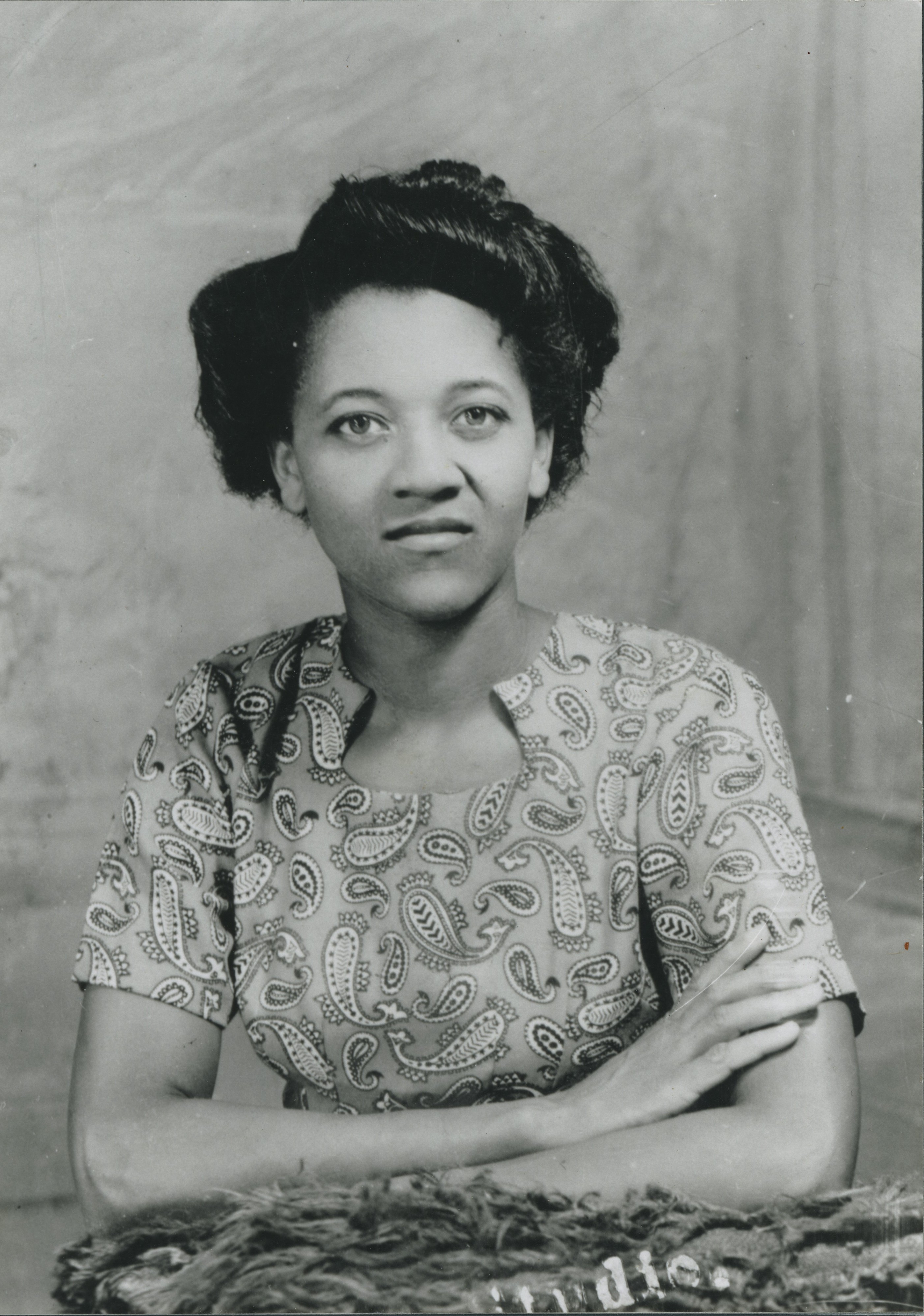 A black and white portrait of a black woman looking directly into the camera. She wears a patterned dress and has her arms folded.
