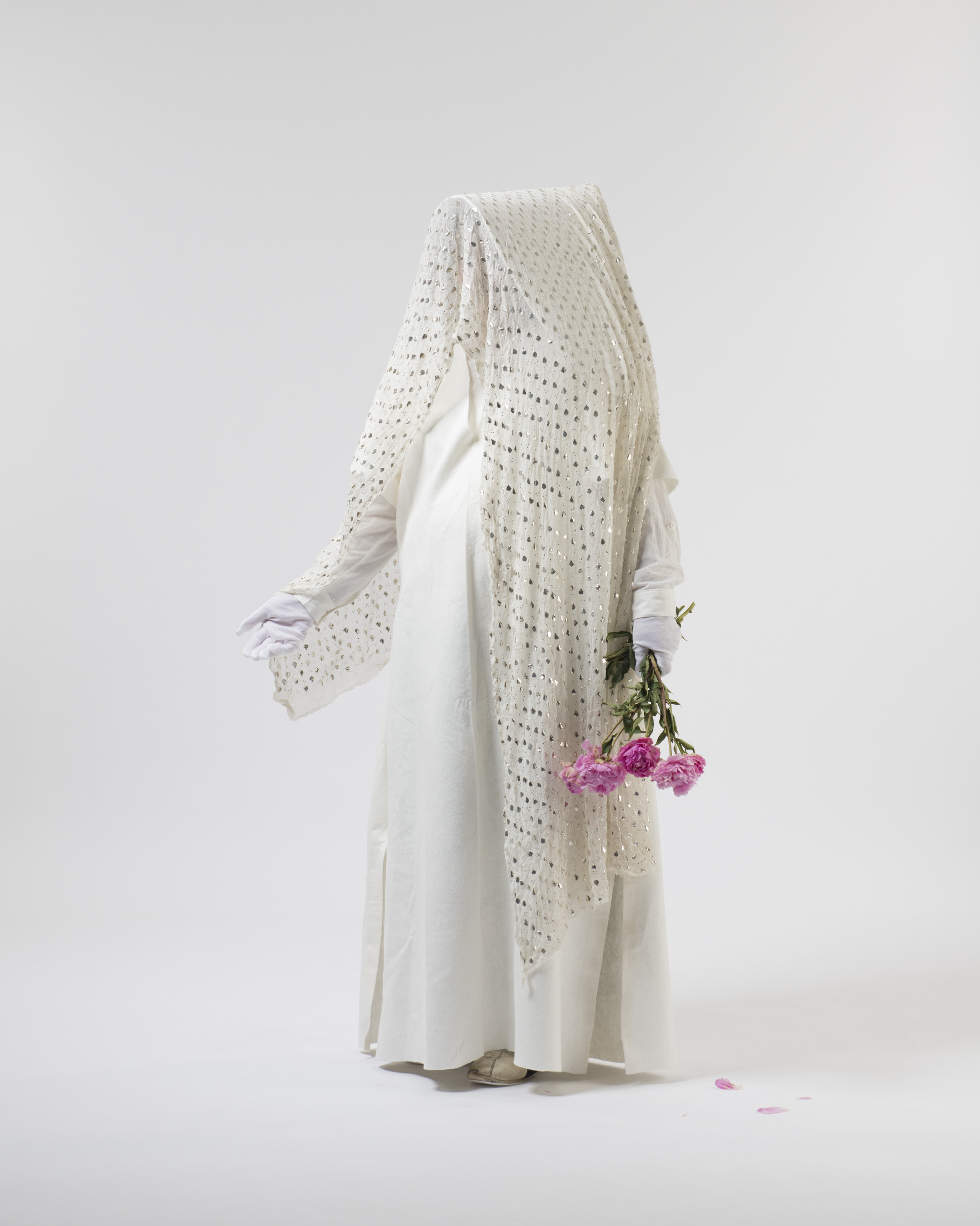 A costumed person against a plain background. The costume is all white, including a dress, gloves and shoes. Over their head is a white scarf with metallic details. They hold a bunch of pink flowers in one hand.