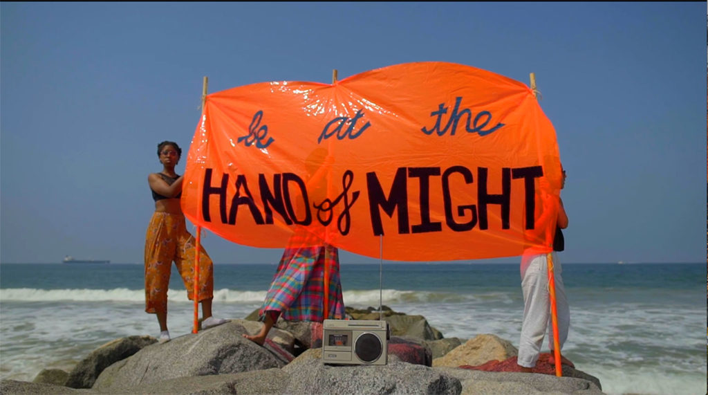 A fabric banner held up by a black woman on a beach. The banner is orange and reads 'be at the hand of might'